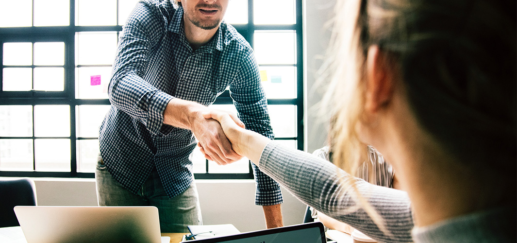 A Smartly Dressed Man in Casual Attire, Firmly Shaking Hands with a Seated Woman in an Office Setting, Symbolising Professional Interaction.